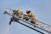 Two firemen on the ladder of the ladder truck spraying water onto a fire nearby.